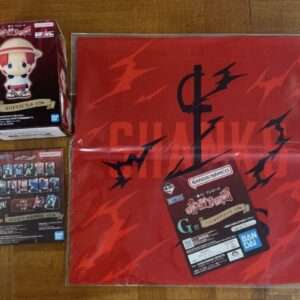 Small chibi figure box on left and a black card with character images on the bottom left, all on a wooden table with a red folder on the right (Bandai Namco items).