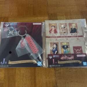 Two sealed anime merchandise packages on a wooden floor: left shows a keychain with red spiral charm; right shows a poster-style sheet of One Piece character art.