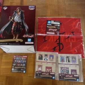 Boxed One Piece figure of Shanks in a long coat and shirt, on a wooden floor beside red packaging with Bandai Namco logos.