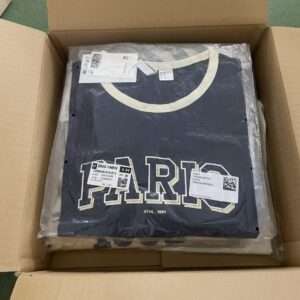 Folded dark T-shirt in clear plastic packaging inside an open cardboard shipping box; cream collar and large white-outlined lettering on the front, plus product tags visible.