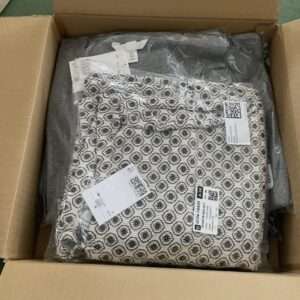 Open cardboard shipping box with a folded patterned garment in clear plastic and attached tags.