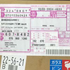 Pink Japanese delivery label stuck to a brown cardboard box, showing multiple tracking numbers, a barcode, and printed text in Japanese.