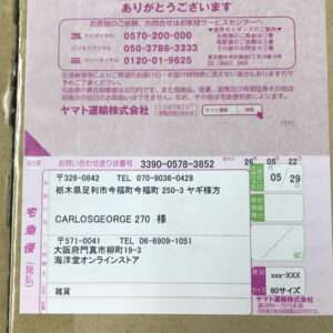 Japanese delivery label on a cardboard box showing recipient name 'CARLOSS GEORGE 270' and address, with sender info, postage marks, and tracking numbers in pink/white forms cover.
