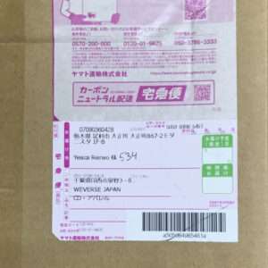 Close-up of a Japanese delivery label on a brown box, showing sender/recipient info, tracking numbers, a barcode, and pink promotional graphics at the top.