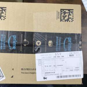 Brown cardboard Amazon box sealed with blue Prime tape, a white shipping label attached showing date, order number, and barcodes