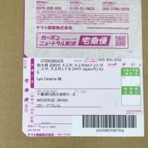 Japanese shipping label on a cardboard box, showing recipient Lya Casana, sender WEVERSE JAPAN, and a barcode with purple sections of the form.