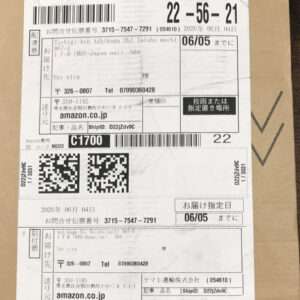 White shipping label on a brown box showing barcodes, QR codes, and Japanese text (Amazon.co.jp tracking and addresses).