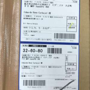 Shipping label on a cardboard box with Japanese text, addresses, tracking numbers, and barcodes visible on blue-bordered sections.