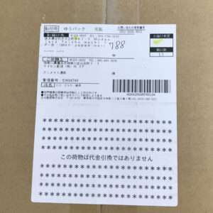 Shipping label with Japanese text and barcode affixed to a brown cardboard box (delivery label viewpoint).