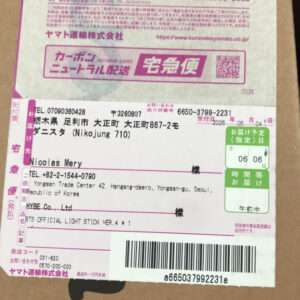Shipping label on a cardboard package with pink accents, barcode at bottom, and a recipient name partially visible as 'Nicolas Mery' (name not identified). Includes tracking numbers and green delivery date box on right side.