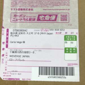 Shipping label on a brown cardboard box with pink and green sections; shows recipient 'Carla Vega' and sender 'WEVERSE JAPAN' with a barcode below.