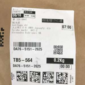 Shipping label on a cardboard box with delivery details: date 06/16, time window 05:00–13:00, recipient Alexandra Rodriguez in Ashikaga, Japan, weight 0.2 kg, tracking numbers, and barcodes/QR codes (Rush Delivery).
