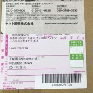 Close-up of a pink Japanese delivery label on a cardboard box, showing recipient name 'Laura Tellez 様', address lines, a barcode at the bottom, and a small green delivery-date box on the right.