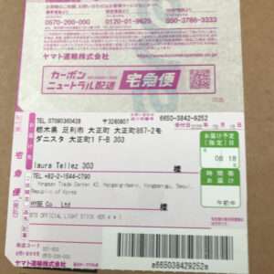 Japanese shipping label on a cardboard box, featuring a barcode, QR code, pink borders, and recipient/sender details in Japanese and English.