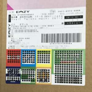 Cardboard box with a white shipping label and barcode, plus Japanese text and a row of colorful grid stickers across the bottom.