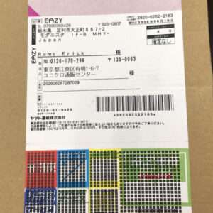 White shipping label with Japanese text, recipient name, and a barcode on a brown cardboard box; colorful grid pattern at the bottom edge suggests branding or packaging design.