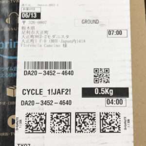 Shipping label on a cardboard box showing destination address, date 06/13, weight 0.5 kg, and a barcode with QR codes nearby.