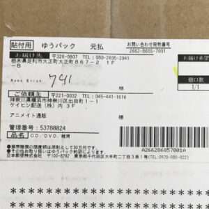 Shipping label affixed to a cardboard box, showing sender and recipient details in Japanese, with a barcode and small black stamp marks.