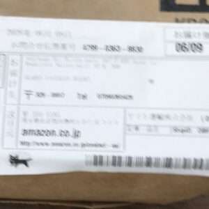 Blurred photo of a cardboard box with a white shipping label showing a barcode and small black logo in the bottom-left corner of the label.