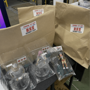Assorted anime figurines in clear plastic cases on a table with brown paper prize bags labeled 雑貨賞 in Japanese sanitary font.