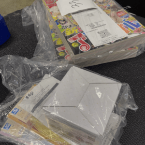 Stack of colorful manga magazines wrapped in clear plastic on a dark carpeted surface, with a white box nearby and receipts on top.