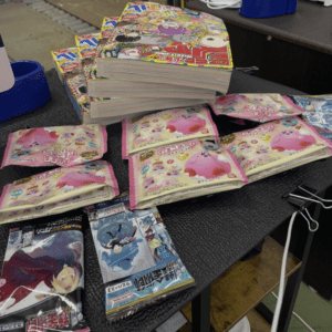 Stack of vibrant manga volumes on a desk, with scattered pink character-themed snack wrappers in front.