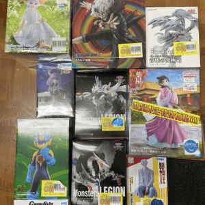 Collection of boxed anime and game figures arranged on a wooden floor, showing colorful character boxes and various brands (Grandista, Monster Legion, etc.).