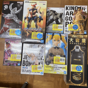 Assorted boxed collectible figures on a wooden floor, featuring Ultraman, a giant Gundam-style model, One Piece art, and a tall Juggler water bottle box in the corner.