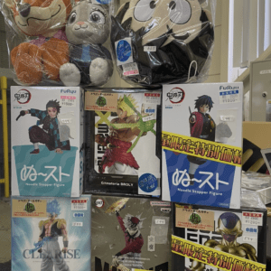 Row of boxed anime figures on a shelf with two large plush toys in clear plastic bags perched on top; store display with price tags visible.