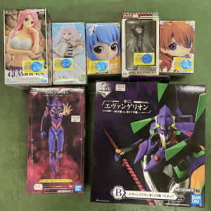 Assortment of boxed anime figures arranged on a green floor, including pink-haired and blue-haired characters among smaller boxes and large Evangelion-model boxes on the bottom row.