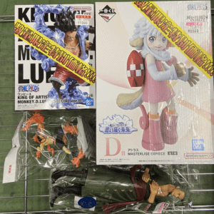 Two large boxed anime figures on a display shelf, with several smaller figures in plastic bags below on a wire rack awaiting sale/collection.