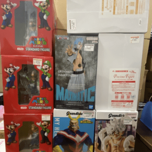 Stack of boxed action figures on a store shelf, including red Super Mario Standard Figure boxes on the left and a Grimmjow MaxMatic box in the center, with Grandista figures at the bottom and large gray boxes in back.