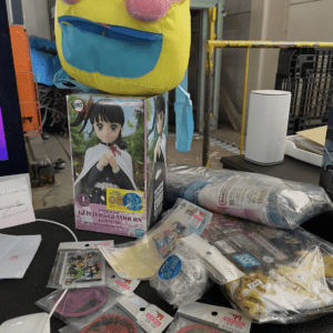 Pink plush Kirby-like character perched on a box featuring a Demon Slayer figure, with anime merch scattered on a desk.