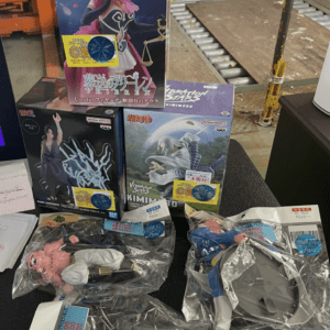 Boxed anime figures and several loose figures in plastic bags displayed on a black table in a warehouse setting.