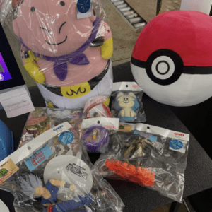 Large pink cartoon plush wrapped in plastic beside a red-and-white Poké Ball plush, with various small packaged figures and toys on a table.