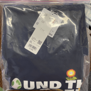 Navy blue folded garment in a sealed plastic bag with attached price tags; size XL visible, plus small decorative graphic (mushroom with a flower) near the bottom edge.