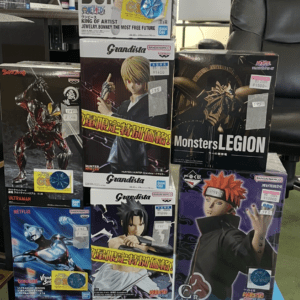 Stack of boxed anime action figures on a display shelf in a shop, with several brands visible (Grandista, Monster Legion, One Piece, Naruto, Ultraman) and price stickers scattered across boxes.