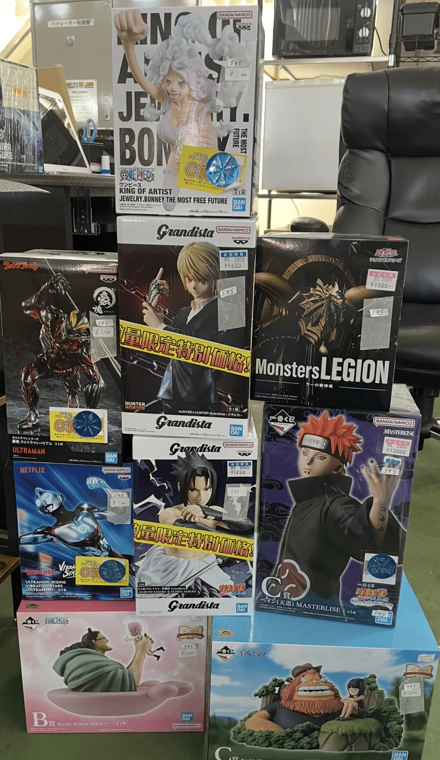 Ver Fotos Recientes 308 - Articulos Japan (post Box Japan) Stack of boxed anime action figures on a display shelf in a shop, with several brands visible (Grandista, Monster Legion, One Piece, Naruto, Ultraman) and price stickers scattered across boxes. Articulos Japan (Post Box Japan)