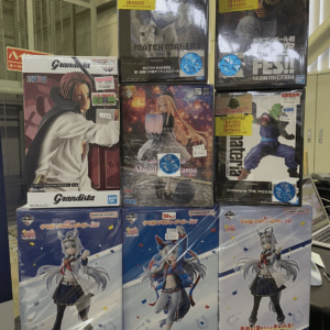 Stacked boxes of Japanese anime figures on a store display, showing character art and price stickers on each box.