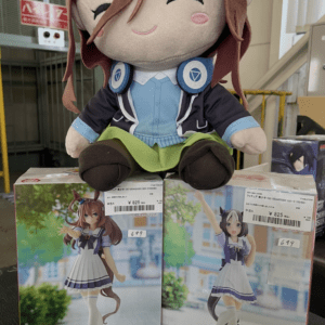 Plush doll with brown hair and blue outfit perched on top of two anime figurine boxes in a store display.