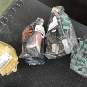 Three wrapped items on a dark surface: a yellow toy on the left, a pair of sneakers in the middle, and a geometric-patterned bag on the right, all in plastic wrap.