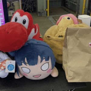 Group of soft plush toys on a desk in a warehouse setting, including a blonde character, a blue-haired round plush, a red dinosaur, and a yellow creature beside a large brown paper bag.
