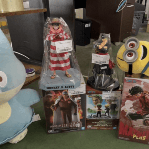 Group of collectible figures and plushies on the floor, including a large blue plush on the left, wrapped figures in the center, and a Minion plush on the right.
