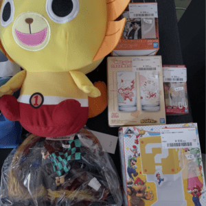 Large yellow plush mascot with orange fins and red shorts surrounded by boxed anime figures on a display table.  Articulos Japan (Post Box Japan)