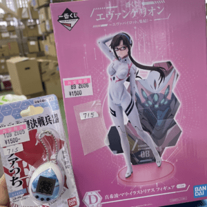Pink Evangelion figure box with a female pilot in a white suit and a large mech illustration on the front, shown in a store setting next to a small keychain package.