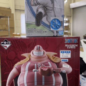 Two stacked boxed anime figures on a store shelf: the top box features a soccer-playing male character (Captain Tsubasa) in a white uniform with a blue swoosh; the bottom box shows a large rotund One Piece character in a red-striped shirt, holding a meat item.