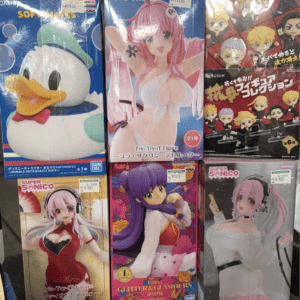 Boxed anime figures on a store shelf: a large Donald Duck/Disney character box on the left, a pink-haired character in a white bikini center, and chibi figure boxes on the right with price tags visible.
