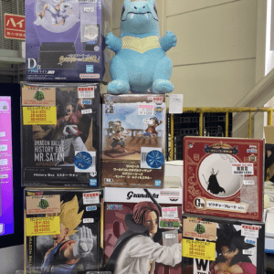 Colorful toy display: large blue Charizard-like figure atop boxes with Dragon Ball figures and plush characters nearby in a store window display.