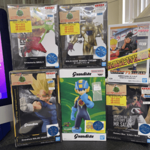 Row of boxed anime figures on a store shelf, including Dragon Ball Grandista boxes featuring Vegeta and Goku and a Naruto box, all in plastic wrap with price stickers visible.