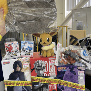A display of anime merchandise: a plush Eevee sits atop red and other boxed figures (Naruto/Boruto) while a gray herringbone hat in a plastic bag is held above, with price tags visible in a shop setting.